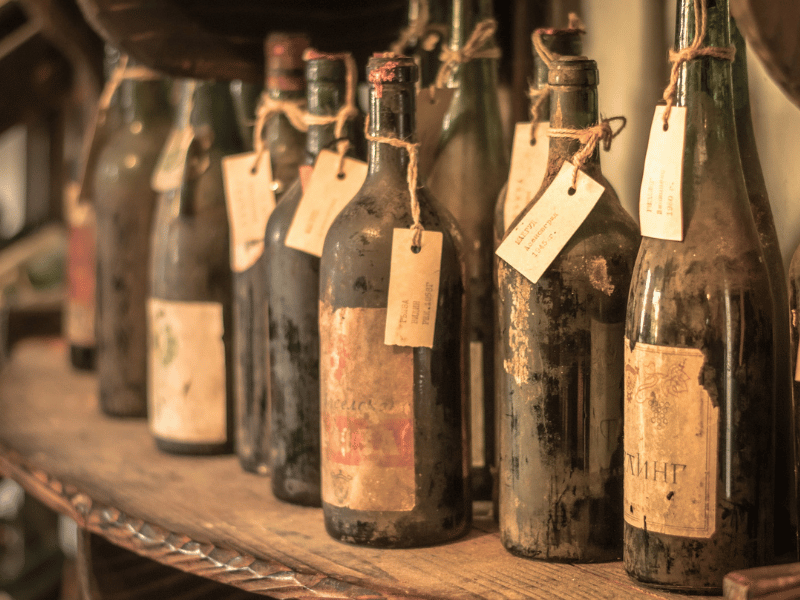 The picture is of several dusty wine bottles sitting on a wooden shelf or table. Some of the bottles have are faded nearly to illegibility. All of the bottles have a separate paper tag tied to the neck with a loop of brown twine. The tags appear to be typewritten.
