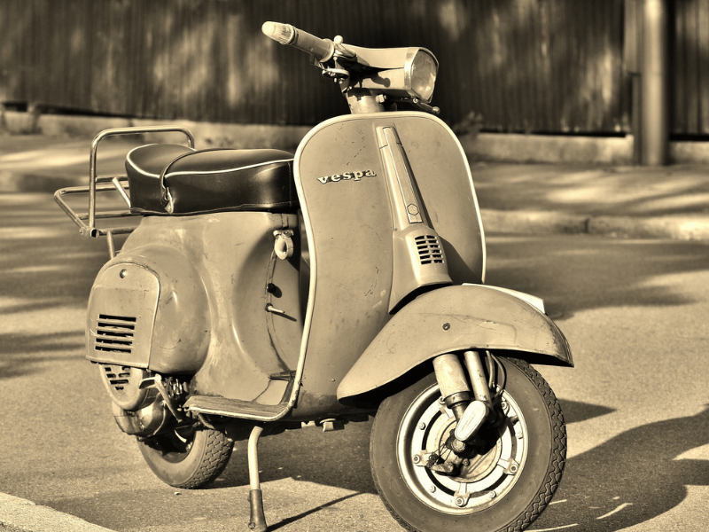 The picture is of a olive drab green Vespa scooter, unoccupied and parked by the curb of a small deserted street. Across from the scooter is a sidewalk and a fence. We can not see very much of the scene around the scooter. The entire picture seems to have a greenish "wash" over it, so that may be the actual color, the picture may have a filter applied to it, or the picture may have a bit of age to it.