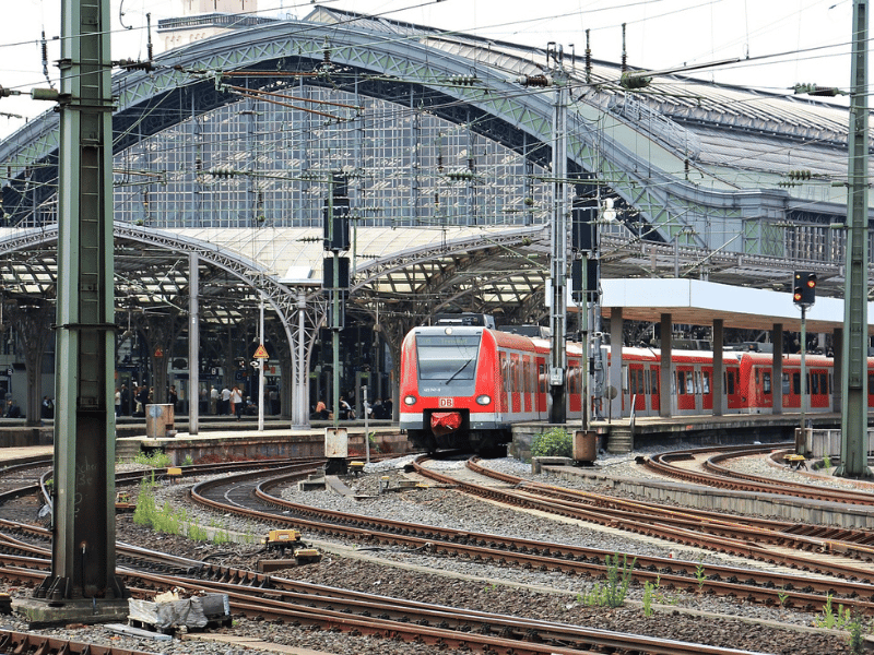 (The picture is of a modern train, red in color, leaving a large metropolitan train station. The station is all grey metal and glass and we can see a bit of the tall buildings beyond it on the left side of the photo. Rails fill the fore and mid-ground -- going into and out of the station and all which-way. Tall electrical towers stand every ten to twenty feet among the rails.)